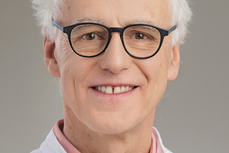 Älterer Mann mit weißem lockigem Haar trägt ein helles Hemd unter einem weißen Kittel und eine Brille