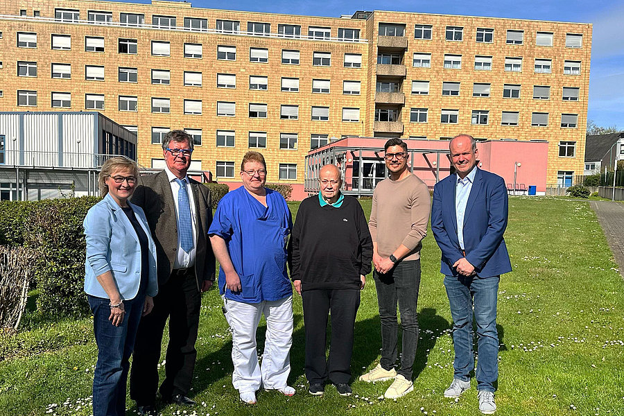 Sechs Personen stehen auf einer Wiese vor einem mehrstöckigen Gebäude unter blauem Himmel