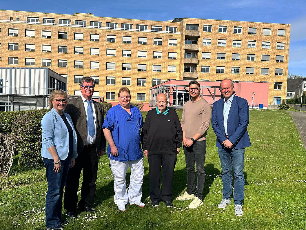 Sechs Personen stehen auf einer Wiese vor einem mehrstöckigen Gebäude unter blauem Himmel