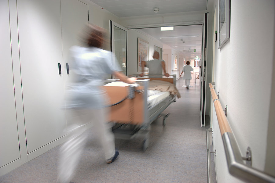 Krankenhausflur mit zwei Personen in weißer Kleidung, die ein Bett schieben, und einer weiteren Person am Ende des Flurs