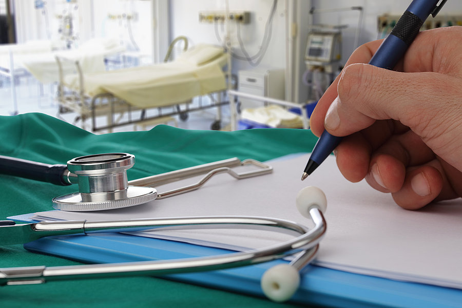 Hand mit Stift schreibt auf einem Klemmbrett neben einem Stethoskop auf einem grünen Tuch in einem Krankenhauszimmer